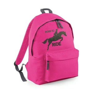 May include: A bright pink backpack with a black graphic of a horse and rider jumping, with the words "BORN TO RIDE". The backpack has a front pocket, a top handle, and adjustable straps. The zippers and handle are gray.
