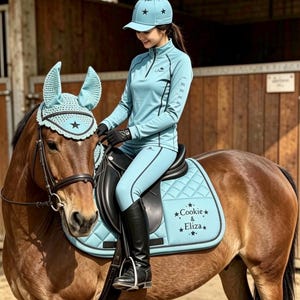 May include: A light blue equestrian set featuring a horse ear bonnet, a quilted saddle pad with the text Cookie & Eliza, a long-sleeved quarter-zip shirt, and a matching baseball cap with a pom-pom, all adorned with black star accents.