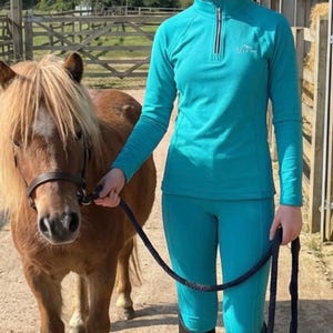 May include: A person in teal riding gear, including a helmet with stars and a white pom-pom, stands beside a brown pony. The person wears a teal top, teal riding leggings, and black riding boots. The pony has a brown bridle.