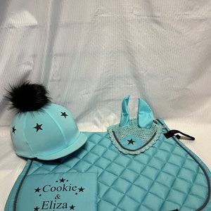 May include: A light blue equestrian set featuring a quilted saddle pad, a matching ear bonnet, and a helmet cover. The saddle pad is embroidered with the text "Cookie & Eliza" surrounded by black stars. Each piece features black star accents.