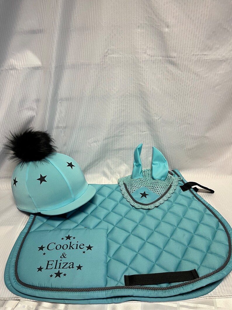 May include: A light blue equestrian set featuring a quilted saddle pad, a matching ear bonnet, and a helmet cover. The saddle pad is embroidered with the text "Cookie & Eliza" surrounded by black stars. Each piece features black star accents.
