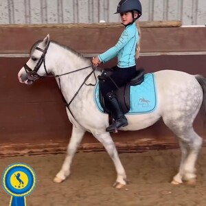 May include: A light gray dapple pony with a black bridle and a black saddle featuring a bright turquoise quilted saddle pad. A rider wears a turquoise long-sleeve shirt, black leggings, black boots, and a dark blue helmet. A blue ribbon icon is visible.