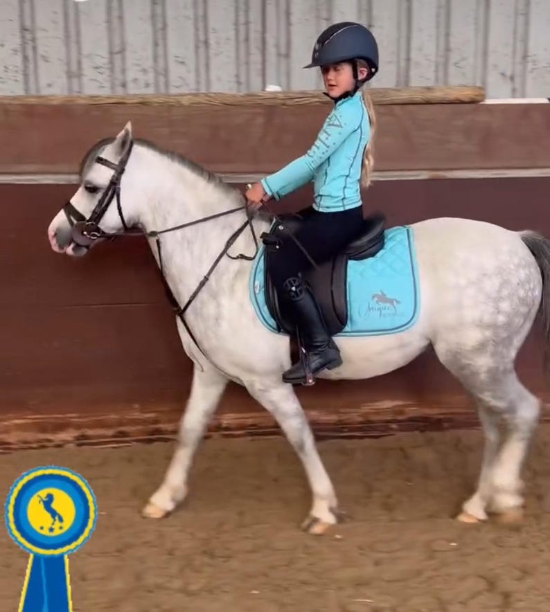 May include: A light gray dapple pony with a black bridle and a black saddle featuring a bright turquoise quilted saddle pad. A rider wears a turquoise long-sleeve shirt, black leggings, black boots, and a dark blue helmet. A blue ribbon icon is visible.