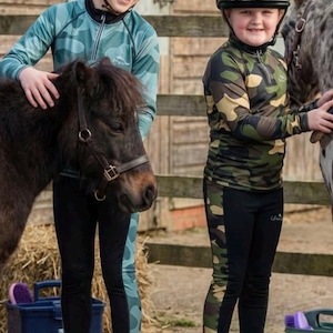 Peut inclure: Deux enfants vêtus d'équipements d'équitation et de casques à motifs camouflage posent avec des poneys. Les casques ont une visière noire et un pompon. Les enfants portent des leggings et des bottes d'équitation noirs. Des bottes de foin et du matériel d'écurie sont visibles.