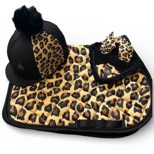 Puede incluir: Un conjunto de equitación que incluye un casco negro con un panel de estampado de leopardo y un pompón negro, una mantilla acolchada con estampado de leopardo y borde negro, y una orejera de ganchillo negra con orejas de leopardo.
