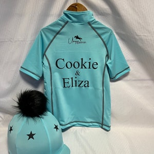 May include: A light blue short-sleeved shirt with a mock neck, featuring the black text "Cookie & Eliza" and a horse logo. Beside it is a matching light blue helmet cover with black stars and a black faux fur pom-pom on top.