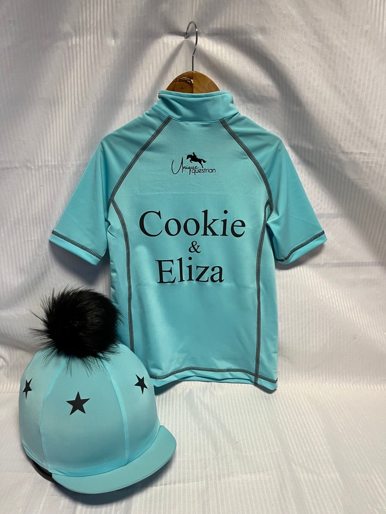 May include: A light blue short-sleeved shirt with a mock neck, featuring the black text "Cookie & Eliza" and a horse logo. Beside it is a matching light blue helmet cover with black stars and a black faux fur pom-pom on top.