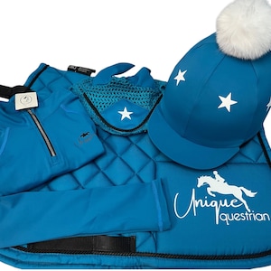 Puede incluir: Un conjunto ecuestre turquesa con una almohadilla de silla de montar, polainas, guantes de equitación y un casco de equitación con un pompón blanco. La almohadilla de silla de montar y el casco tienen estrellas blancas y la almohadilla de silla de montar tiene el texto "Unique Equestrian" en blanco.
