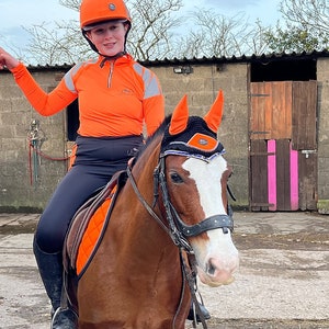 May include: A person wearing an orange riding outfit and helmet is riding a brown and white horse. The horse is wearing a black bridle and a black and orange fly mask.