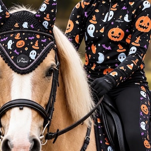 May include: A black equestrian set featuring a Halloween pattern of orange pumpkins, white ghosts, purple bats, and orange witch hats. The set includes a long-sleeved quarter-zip shirt, black leggings, a matching horse ear bonnet, and a helmet cover.