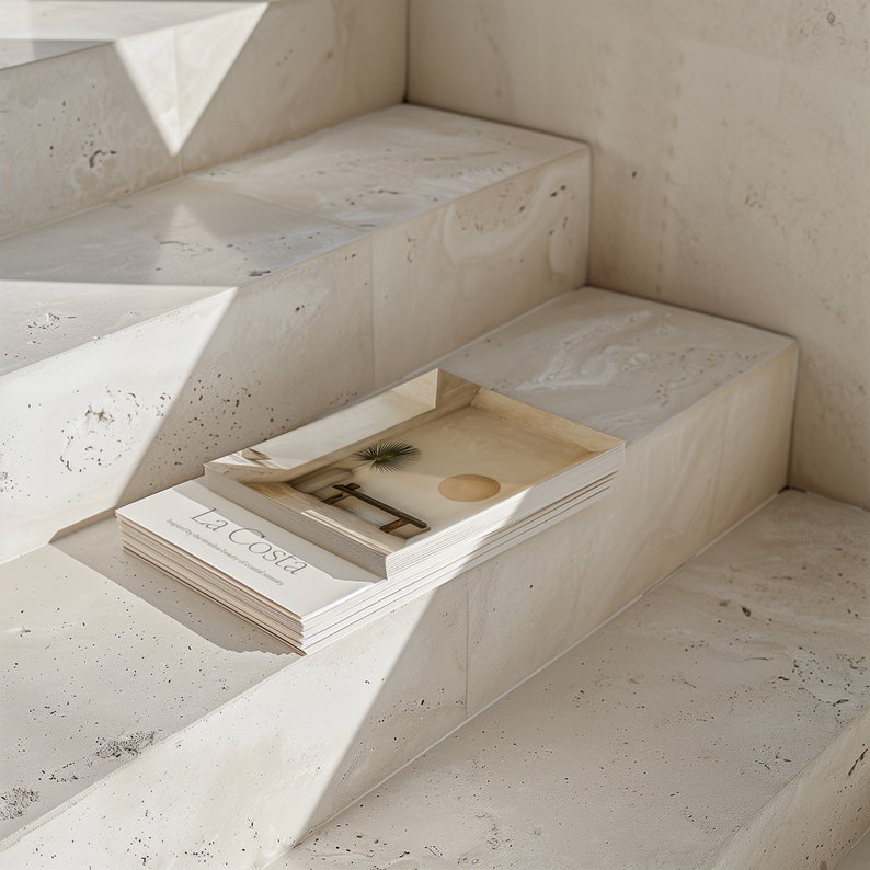 May include: A book titled "La Costa" with a minimalist design featuring a palm tree and a sun on the cover, resting on a set of concrete steps.