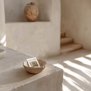May include: A small, beige, textured bowl with a white card inside. The card has the text "La Costa" printed on it. The bowl is sitting on a beige, textured surface.