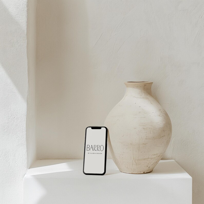 May include: A black smartphone with a white screen displaying the text "BARRO by ALICIA DESIGN" is sitting on a white surface next to a large, off-white ceramic vase.