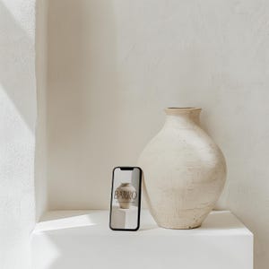 May include: A white ceramic vase with a textured surface sits on a white shelf next to a black smartphone displaying a website for a pottery studio called "BARRO".