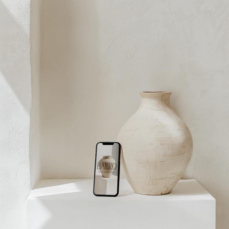 May include: A white ceramic vase with a textured surface sits on a white shelf next to a black smartphone displaying a website for a pottery studio called "BARRO".
