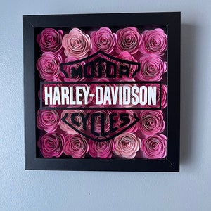 May include: A framed black and white Harley-Davidson logo surrounded by pink paper flowers.