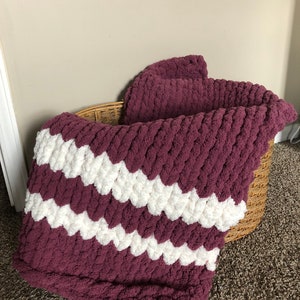 May include: A chunky knit throw blanket with a burgundy and white chevron pattern. The blanket is folded and placed in a wicker basket.