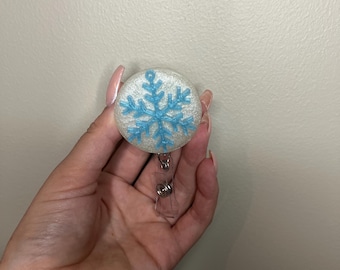 May include: A round, white badge reel with a light blue snowflake design. The snowflake is centered on the badge reel. The badge reel has a clear plastic retractable cord and a metal clip.