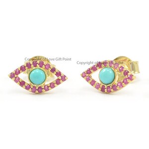 May include: Gold earrings with a turquoise evil eye design surrounded by pink stones.