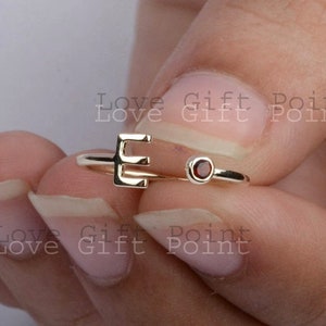 Puede incluir: Anillo de oro con la letra "E" y una pequeña piedra preciosa roja. El anillo está en un dedo.