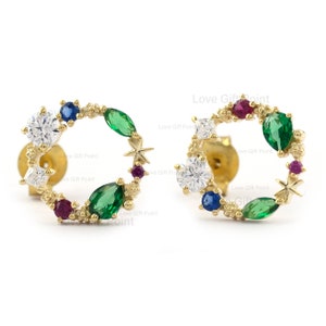 May include: A pair of gold earrings with a circular design featuring a variety of colorful gemstones, including green, blue, red, and white. The earrings are set with small stars and have a delicate, feminine look.