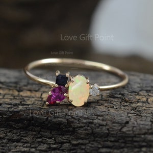 Op de afbeelding: Een delicate gouden ring met een ovale opaal, omringd door kleinere edelstenen in tinten roze, rood en blauw. De ring ligt op een houten oppervlak.