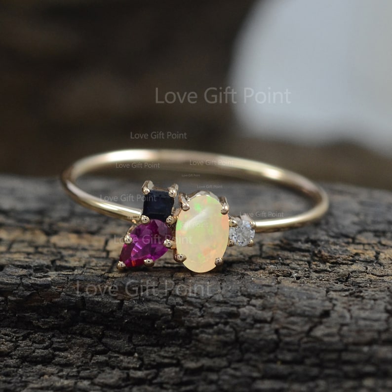 Op de afbeelding: Een delicate gouden ring met een ovale opaal, omringd door kleinere edelstenen in tinten roze, rood en blauw. De ring ligt op een houten oppervlak.