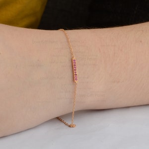 Op de afbeelding: Een gouden kettingarmband met een dunne staafbedel bezet met roze edelstenen. De staafbedel heeft een delicaat, minimalistisch ontwerp.