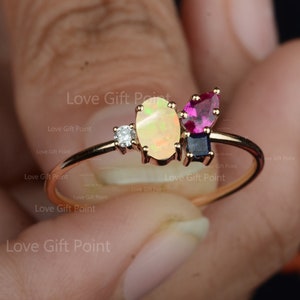Op de afbeelding: Een gouden ring met een witte opaal, een roze edelsteen en een blauwe edelsteen. De ring is bezet met kleine diamanten.