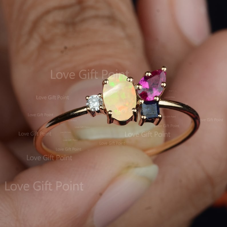 Op de afbeelding: Een gouden ring met een witte opaal, een roze edelsteen en een blauwe edelsteen. De ring is bezet met kleine diamanten.