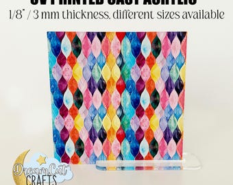May include: A colorful abstract design printed on a 1/8 inch thick clear acrylic sheet. The design features a repeating pattern of overlapping teardrop shapes in shades of blue, green, yellow, pink, purple, and red. The acrylic sheet is standing on a clear acrylic stand.