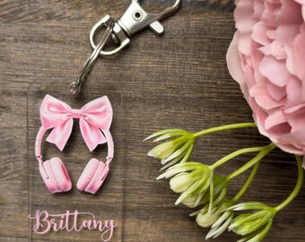 Puede incluir: Un llavero de acrílico transparente con un lazo rosa y auriculares rosas. El llavero tiene el nombre "Brittany" impreso en él.