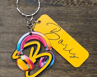 Puede incluir: Un llavero amarillo y negro con una letra B estilizada y auriculares. El llavero tiene una etiqueta amarilla con el nombre "Baris" escrito en negro.