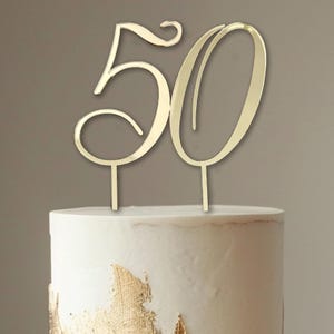 Puede incluir: Topper para pastel de acrílico dorado espejado en forma del número 50. El número 5 está estilizado con un toque de fantasía. El topper está sobre un pastel blanco.