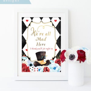 Può includere: Un cartello incorniciato bianco con uno sfondo a scacchi bianco e nero e cuori rossi. Il cartello dice "We're all Mad Here I think you'll fit right in". C'è un cappello a cilindro nero con un'arrangiamento di fiori rossi e bianchi sotto.