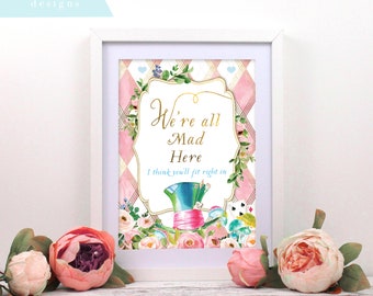 May include: A white framed print with a pink and white checkered background. The print features a gold frame with the text "We're all Mad Here I think you'll fit right in". The image also includes a whimsical illustration of the Mad Hatter's hat, flowers, and playing cards.