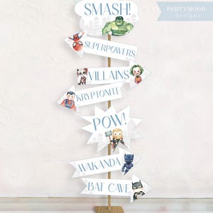 Puede incluir: Un conjunto de decoraciones para fiestas con tem&aacute;tica de superh&eacute;roes con flechas que apuntan a diferentes ubicaciones. Las flechas est&aacute;n decoradas con im&aacute;genes de superh&eacute;roes y villanos populares, incluidos Superman, Batman, Spiderman, Hulk, Thor, Black Panther y el Joker. El texto en las flechas dice "Superpoderes", "Villanos", "Kryptonita", "Pow", "Wakanda" y "Bat Cave".