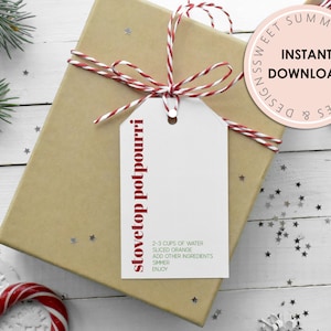 Puede incluir: Una etiqueta de regalo blanca con un cordel rojo y blanco atado alrededor. La etiqueta tiene el texto "stovetop potpourri" escrito verticalmente en letras rojas. La etiqueta también incluye instrucciones para hacer potpourri para estufa, incluyendo el uso de 2-3 tazas de agua, naranja en rodajas y otros ingredientes. La etiqueta está atada a una caja de regalo marrón con un fondo blanco.