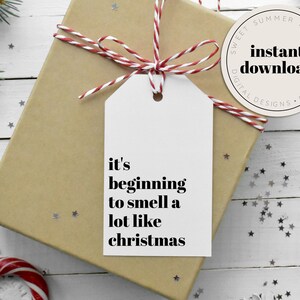 Puede incluir: Una etiqueta de regalo blanca con texto negro que dice "it's beginning to smell a lot like christmas". La etiqueta está atada con una cuerda roja y blanca a una caja de regalo marrón.