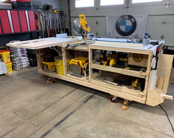 May include: A wooden mobile workbench with a yellow and black power saw, a yellow sander, and other tools. The workbench has wheels and a fold-down side table.