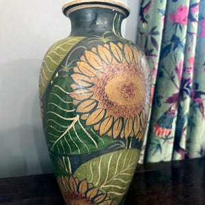 May include: A ceramic vase with a dark background featuring hand-painted yellow sunflowers and large green leaves. The vessel has a narrow neck, a flared rim, and a rounded body, showcasing a detailed botanical pattern with textured brushwork.