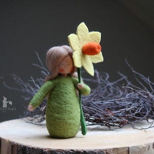 Op de afbeelding: Een vilten pop met lang blond haar, die een groene jurk draagt en een gele vilten narcis met een oranje hartje vasthoudt.