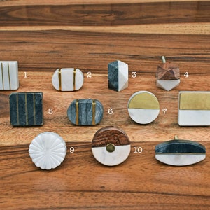 May include: Eleven different styles of marble and wood cabinet knobs with gold accents. The knobs are arranged in three rows on a wooden surface.