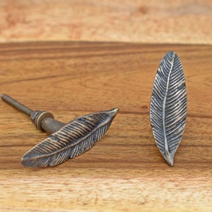 May include: Two metal leaf-shaped cabinet knobs. One knob is attached to a screw, the other is not. Both knobs are a dark bronze color.