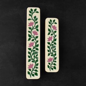 May include: Two ivory-colored rectangular decorative pieces with a floral design. The design features pink flowers and green leaves on a winding vine. The pieces are set against a dark background.