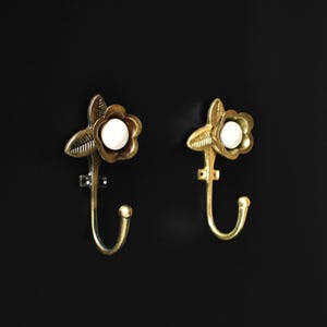 May include: Two gold-toned flower-shaped hooks with white centres. The hooks are mounted on a black surface.
