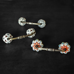 May include: Three ceramic drawer pulls with floral designs. The pulls are white with blue, orange, and green accents. The pulls are attached to a metal base.