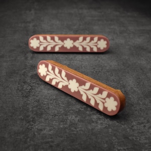 May include: Two pink wooden drawer pulls with a white floral design. The pulls are oval shaped and have a slightly rounded edge.