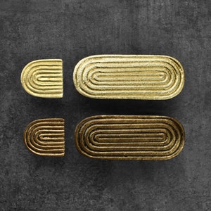 Può includere: Due set di maniglie per cassetti in metallo dorato. Ogni set include una piccola maniglia a semicerchio e una maniglia ovale più grande. Le maniglie hanno un design a cerchi concentrici.