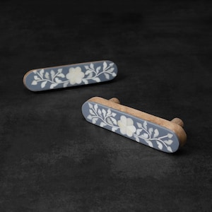 May include: Two grey and wood drawer pulls with a floral design. The pulls are made of wood with a grey painted surface and white floral inlays.